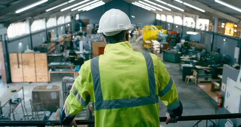Responsable d'exploitation, de dos, en gilet haute-visibilité et casque, observant son atelier depuis une passerelle — le terrain (gemba), là où se crée la valeur.