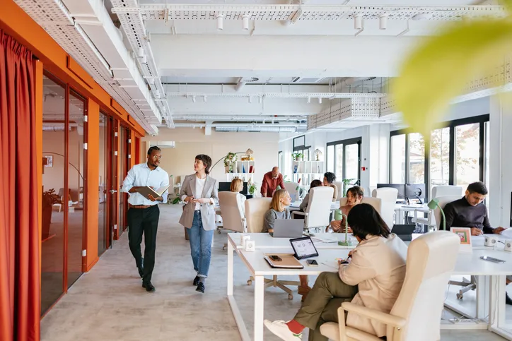 Open space lumineux : deux collaborateurs échangent en marchant tandis qu'une équipe travaille aux postes de travail.